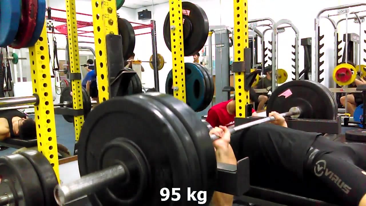 這張照片是一名男子在健身房做臥推運動。他正在舉起 95 公斤的槓鈴，臉上展現出專注的神情。