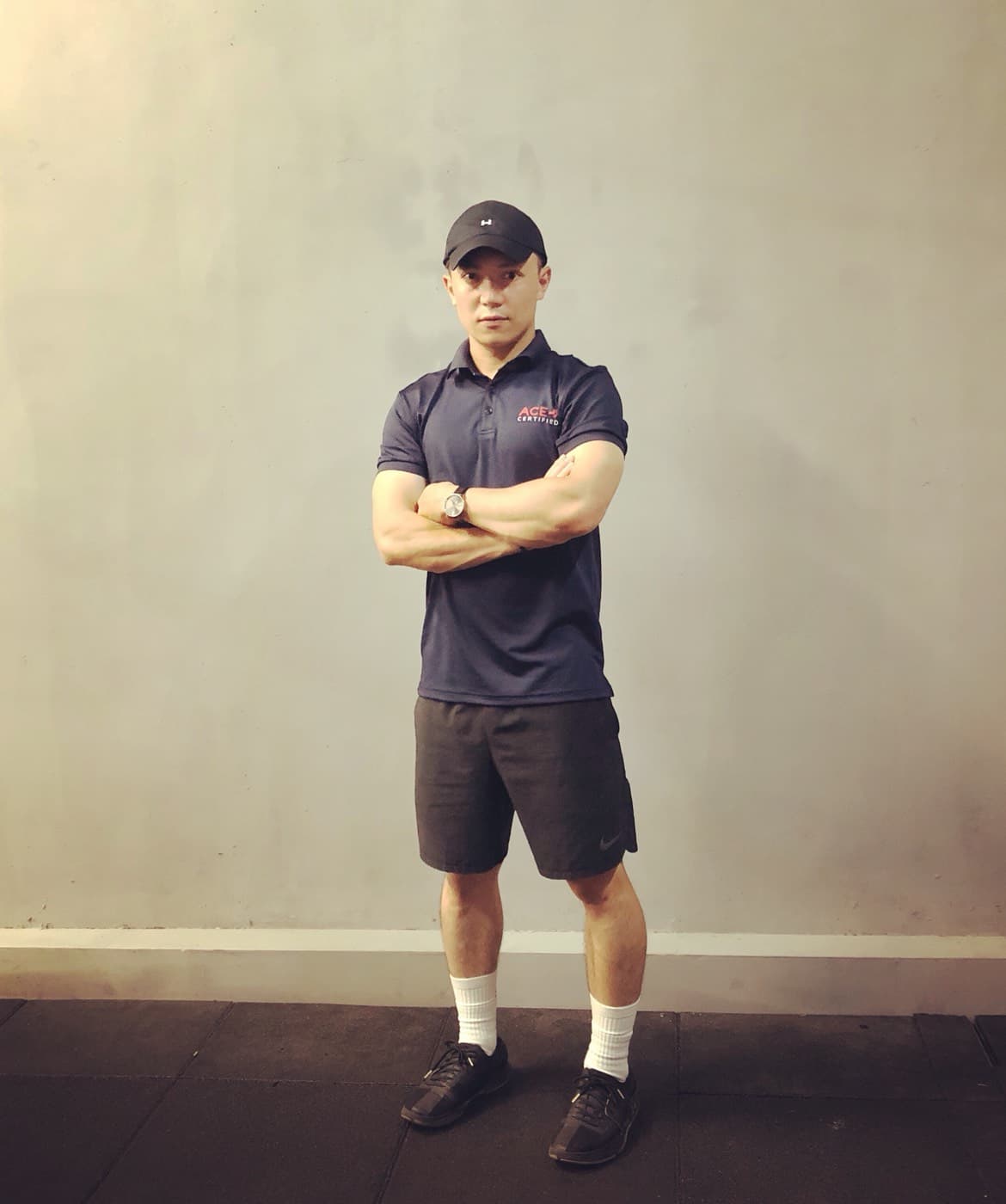 這張照片是一個穿著黑色運動服的男性，他站在一個灰色牆壁的房間裡。他正在看著照相機，並雙手交叉在胸前。他看起來很有自信。