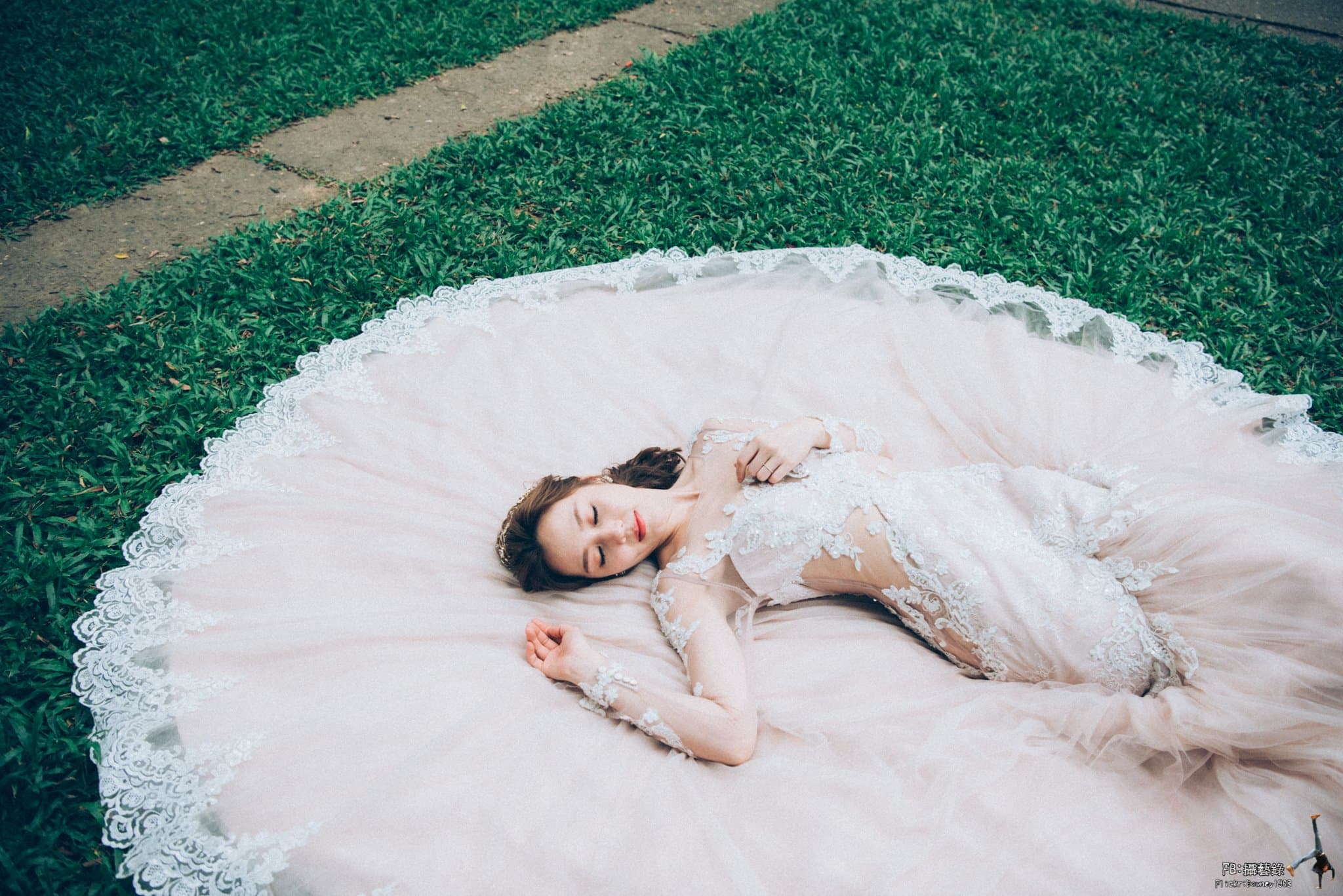 這張「婚紗照」的照片是一個女人躺在草地上，她穿著一件蕾絲婚紗，她的頭髮散落在肩上，她閉著眼睛，似乎在睡覺。