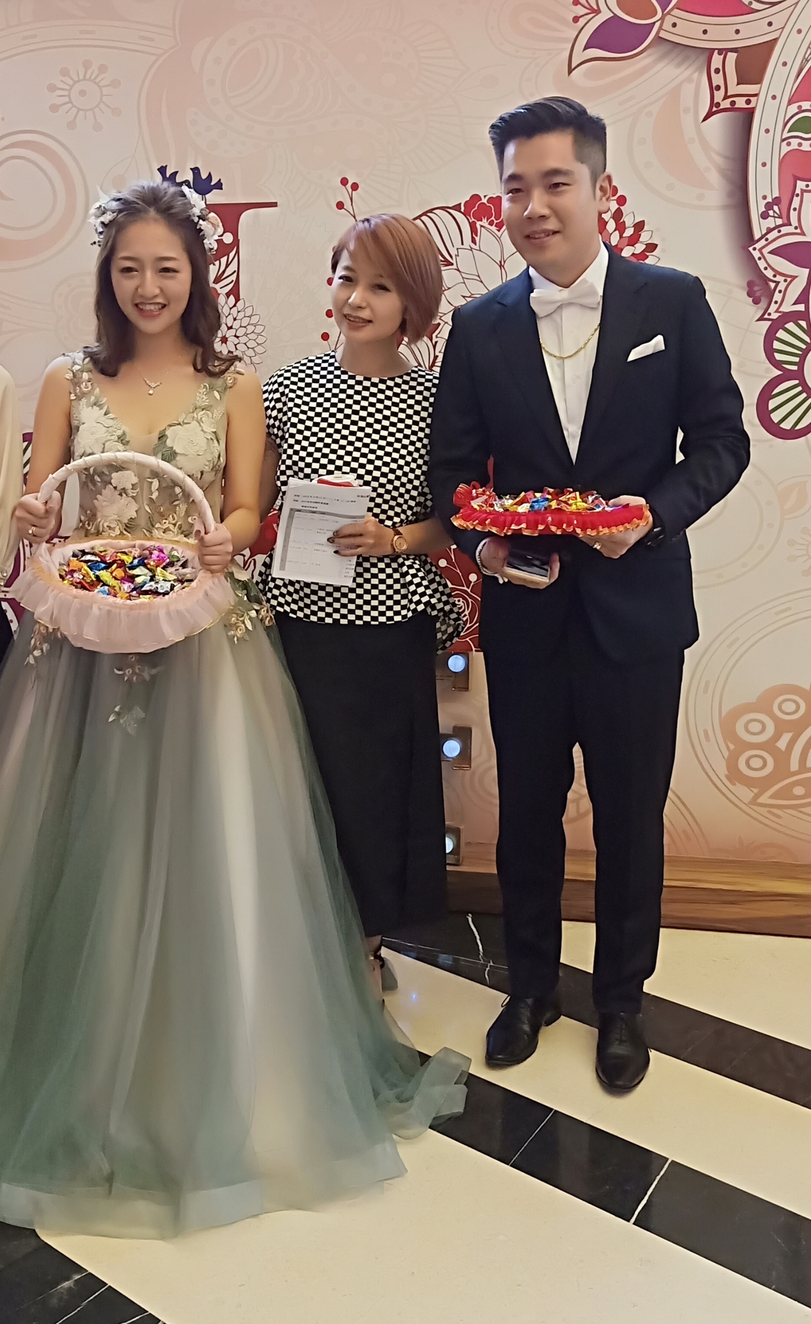 這張婚禮照片是三個朋友，他們穿著正式服裝，正在慶祝婚禮。其中一個女性穿著綠色的禮服，另一個女性穿著黑色格子裙，而男性則穿著深色西裝。他們都拿著禮物，看起來很開心。