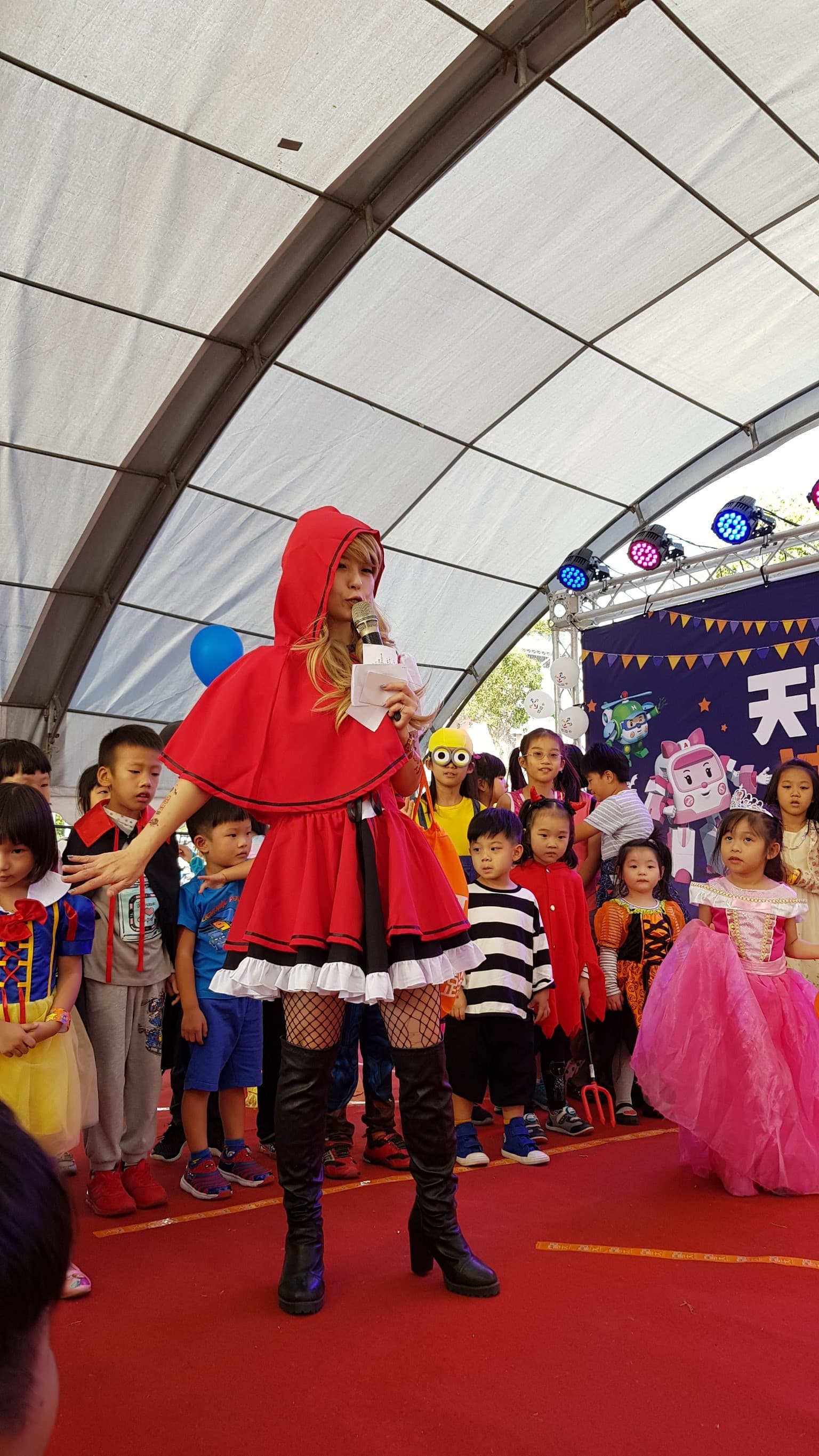 這張「兒童遊行」的照片是一群小朋友在舞臺上表演，其中一個穿著紅色斗篷的女孩子站在舞臺中央，她手裡拿著麥克風，正在唱歌。