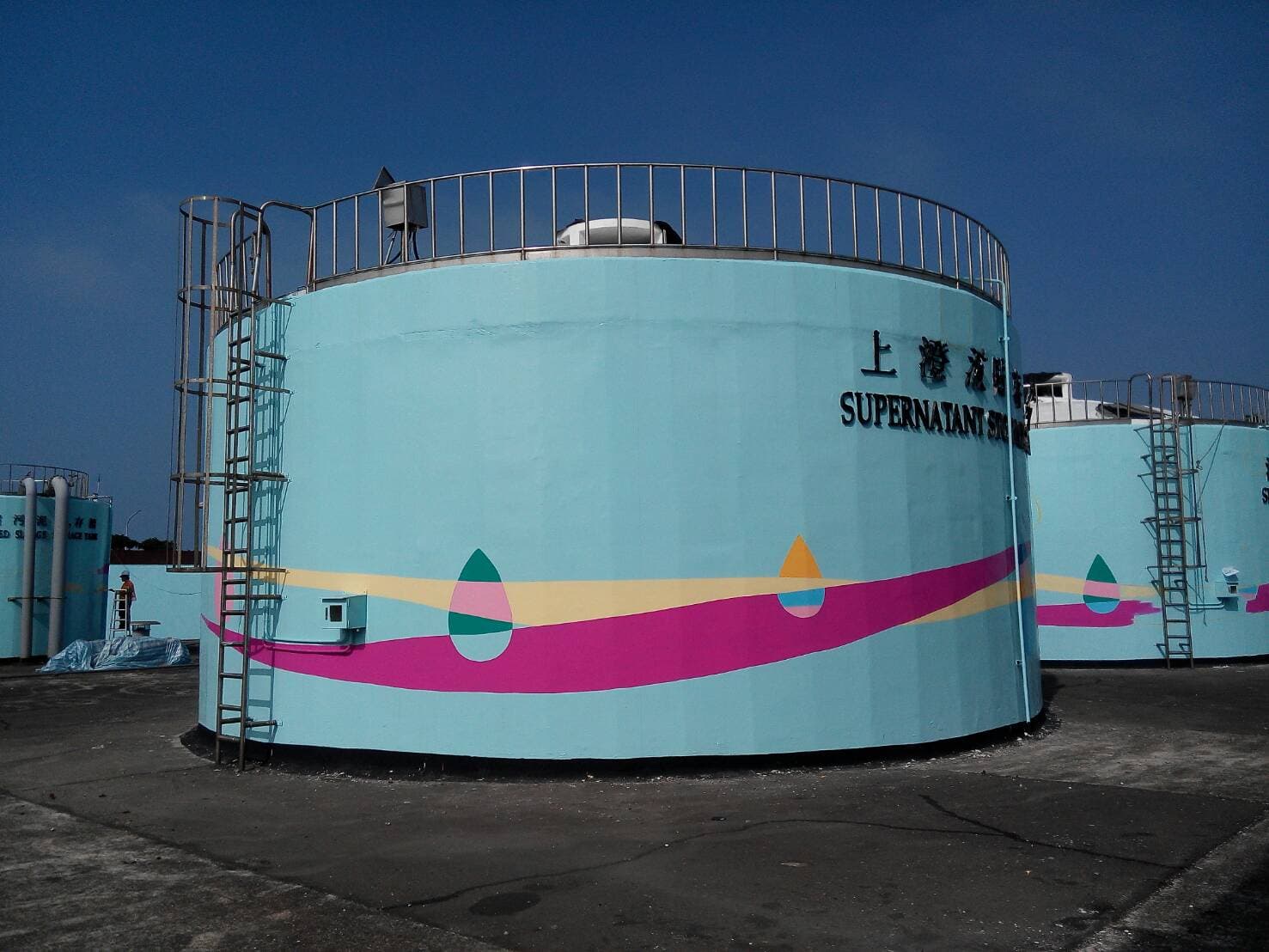 這張「大型儲水槽」的照片是藍色圓柱形的水槽，上面有粉紅色的條紋和三個彩色水滴，水槽旁有樓梯，水槽上寫著「上清液槽」的字樣。