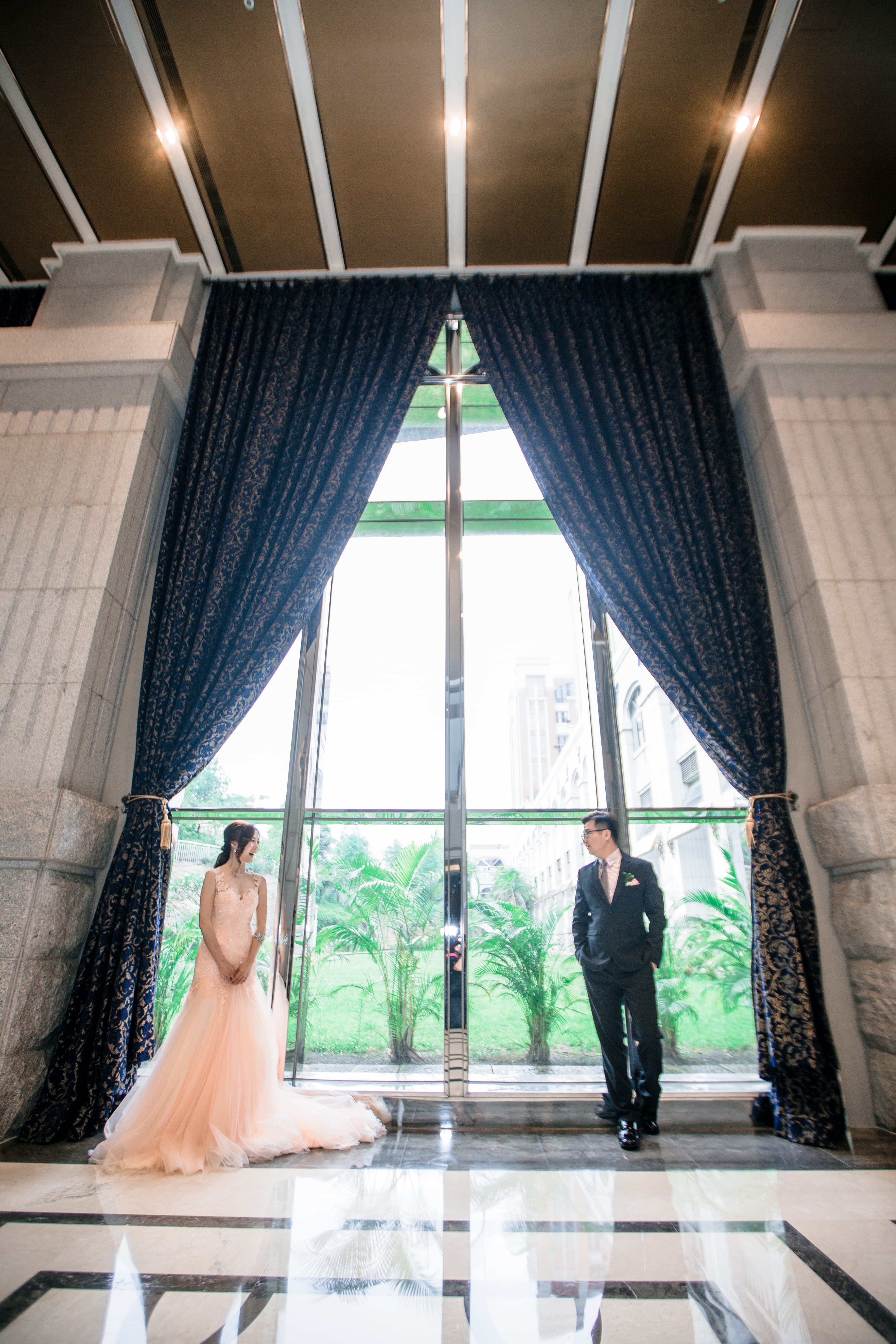 這張照片是一對新人站在窗戶前，他們穿著婚紗和西裝，背景是綠色的植物和城市風景。