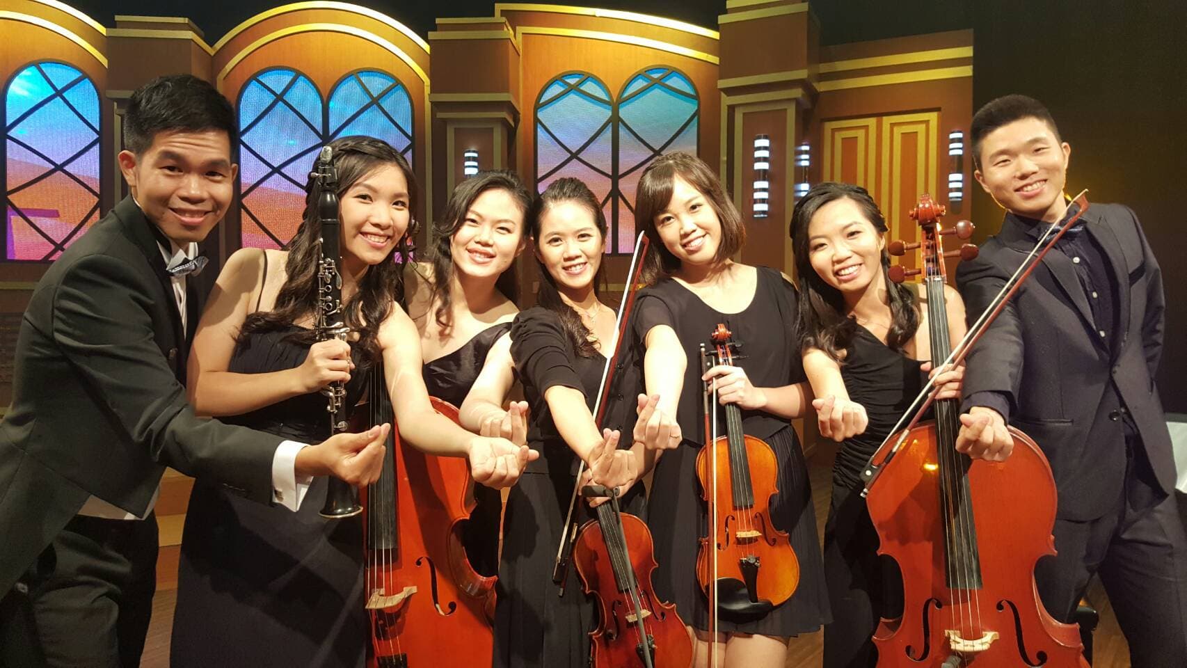 這張照片是一個音樂表演，六位音樂家站在舞台上，其中一位演奏單簧管，兩位演奏小提琴，兩位演奏大提琴，而另一位則手拿一根小提琴。他們都穿著正式的服裝，顯得非常專業。