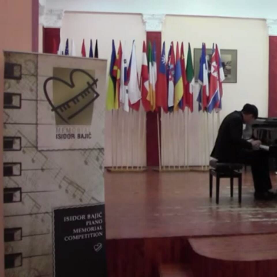 這張照片是一個鋼琴比賽的舞臺，照片最重要的是舞臺上的鋼琴家正在演奏。他穿著黑色西裝，坐在鋼琴前，專注地彈奏著。