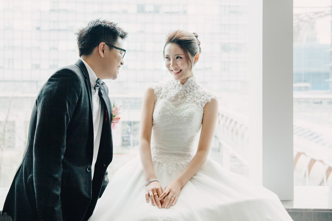 這張婚紗照是一對新人在窗邊互相凝視，新郎穿著黑色西裝，新娘穿著白色婚紗，他們都面帶笑容，看起來非常幸福。