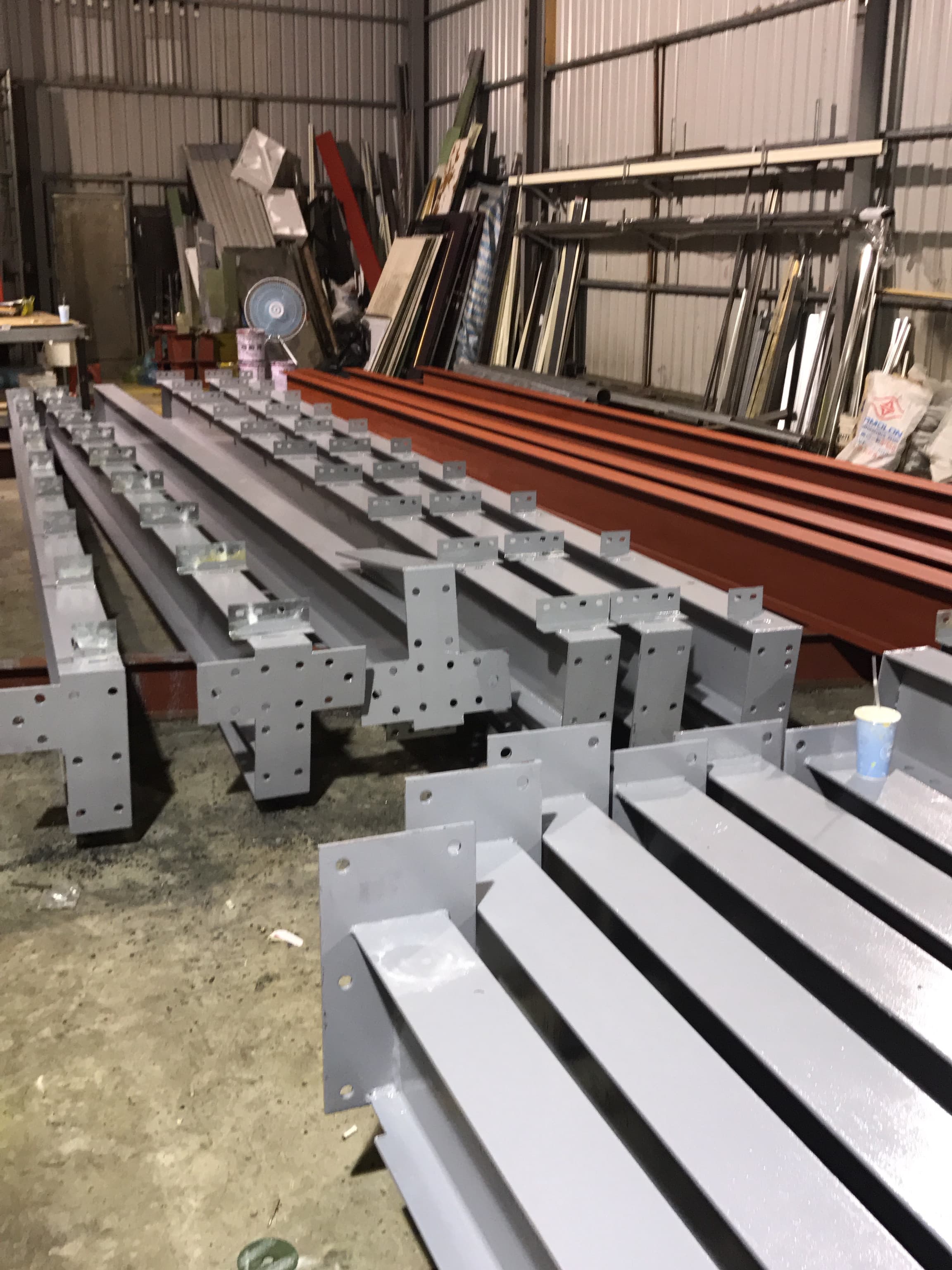 1) 照片顯示一間工廠內，地面上擺放著許多鋼製樑柱，這些樑柱為灰白色，表面看起來是經過油漆處理，照片的背景則是一些雜亂的建築材料