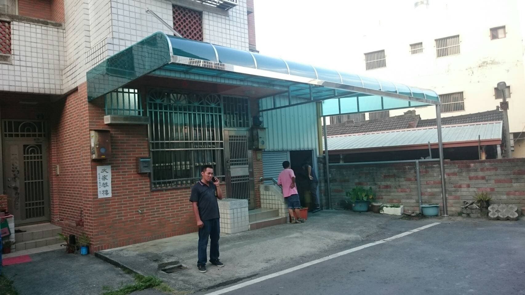 這張「房屋」的照片是 一棟住宅的側面，房屋上有個透明的遮雨棚，遮雨棚下方有一個男子正在看手機。