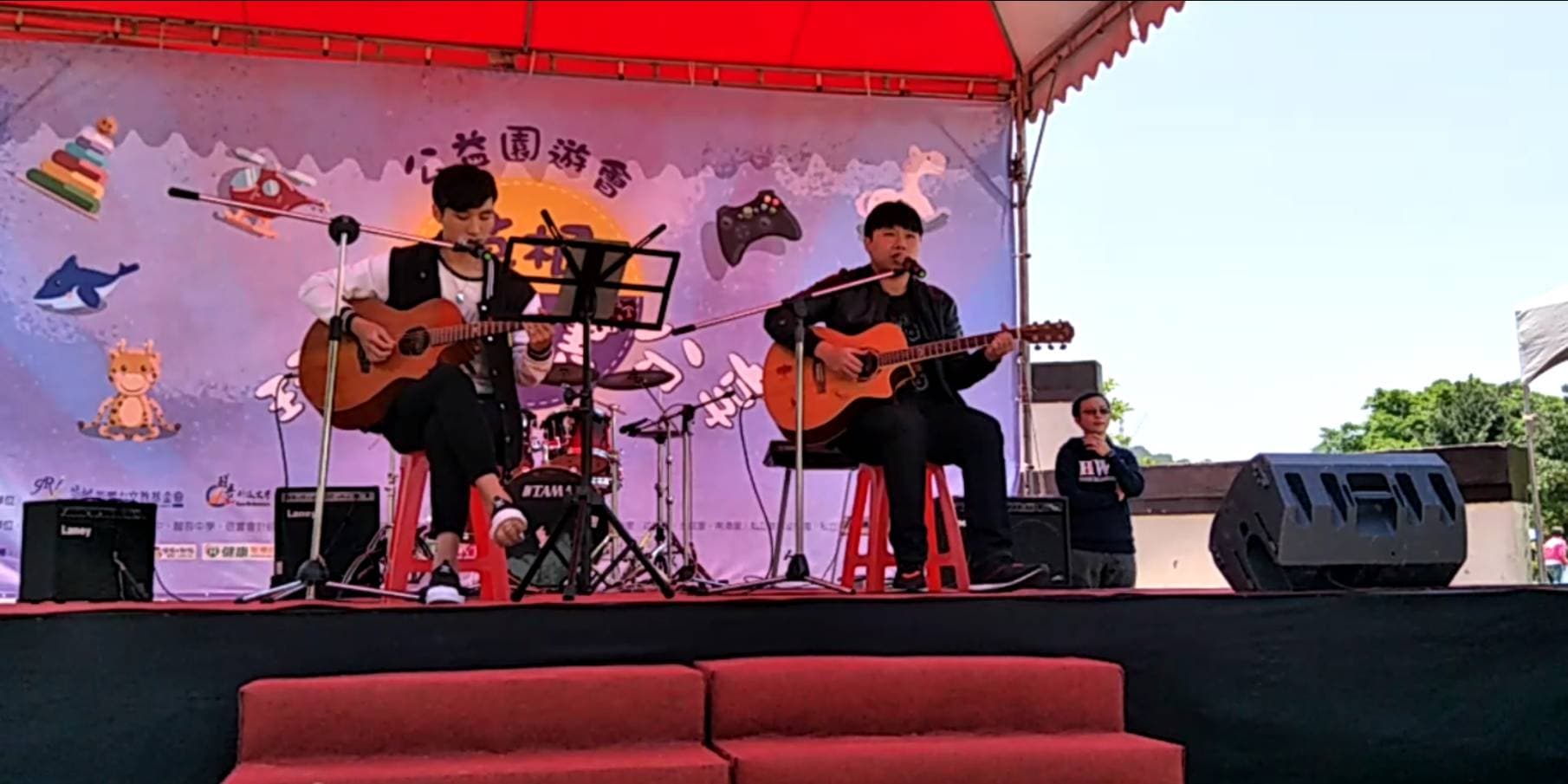 這張「戶外音樂表演」的照片是兩個年輕人正在用吉他唱歌，舞臺上擺著音響和麥克風，背景是紅色的棚架，畫面生動活潑。