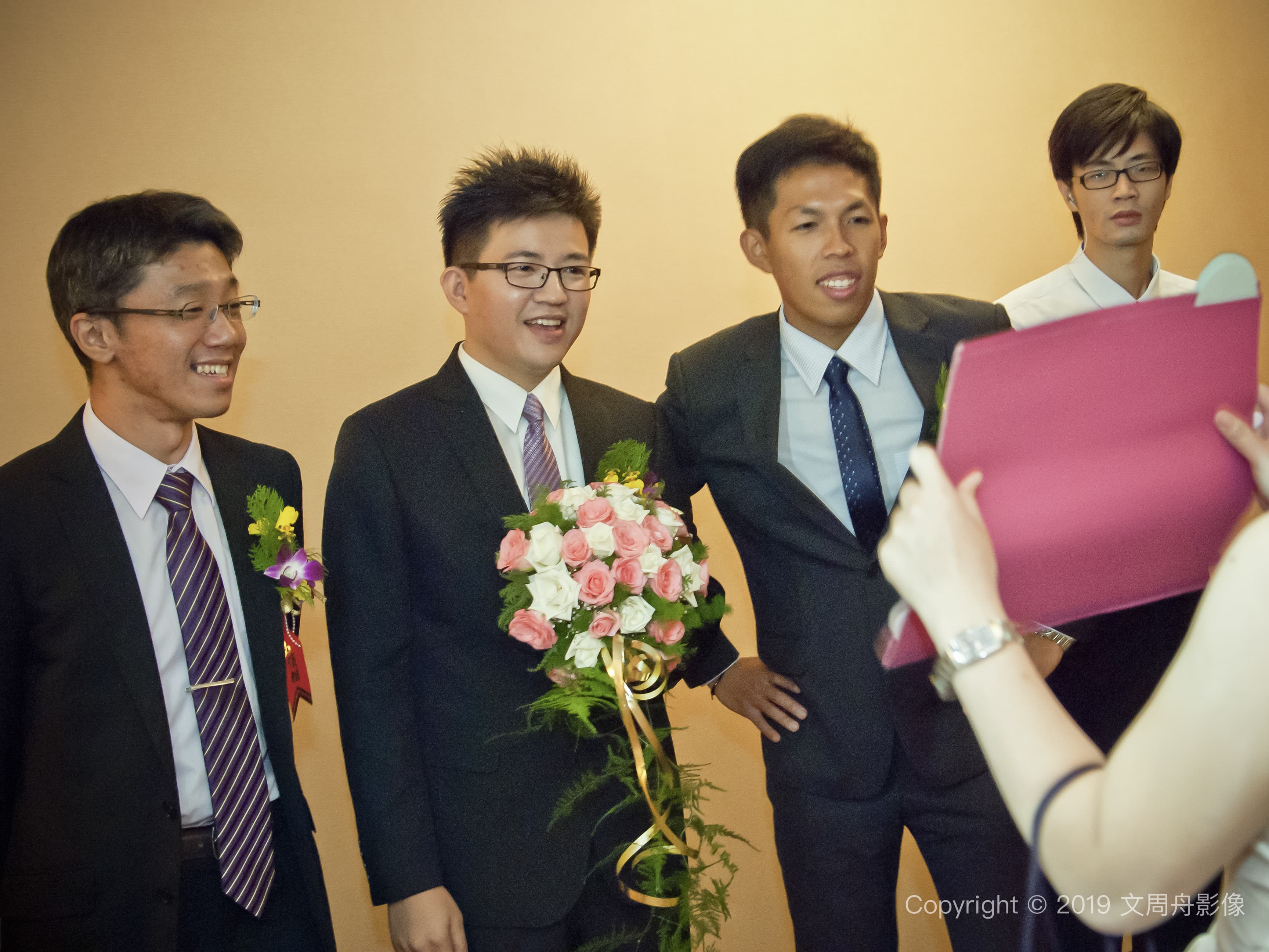 這張照片是四個男人在婚禮上，其中一個穿著白色禮服，站在中間，他正拿著一把鮮豔的玫瑰花。