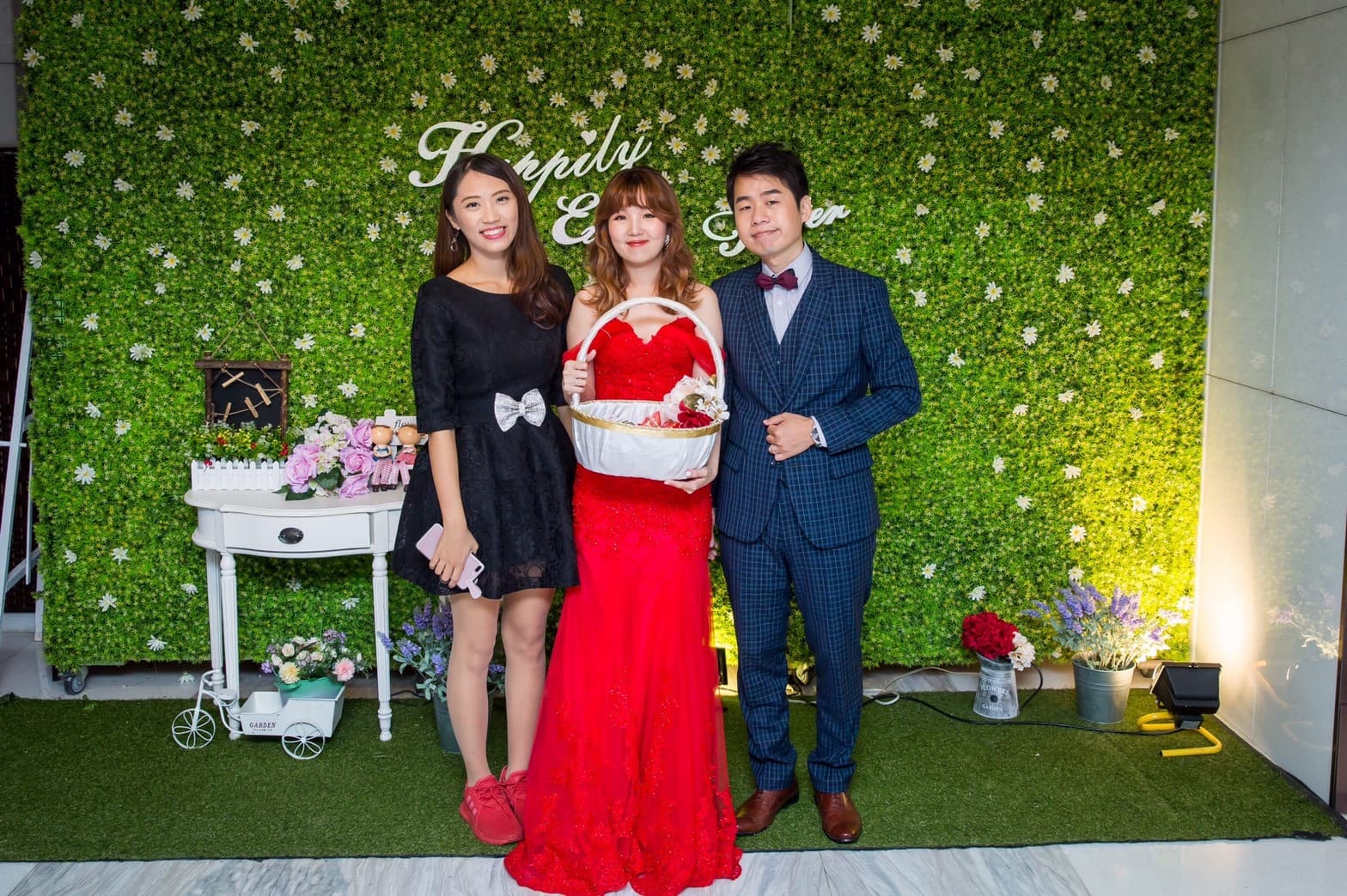 這張婚禮照片是三個人站在綠色背景牆前，中間的女生穿著紅色禮服，旁邊站著穿著黑色洋裝的女生和穿著西裝的男生。