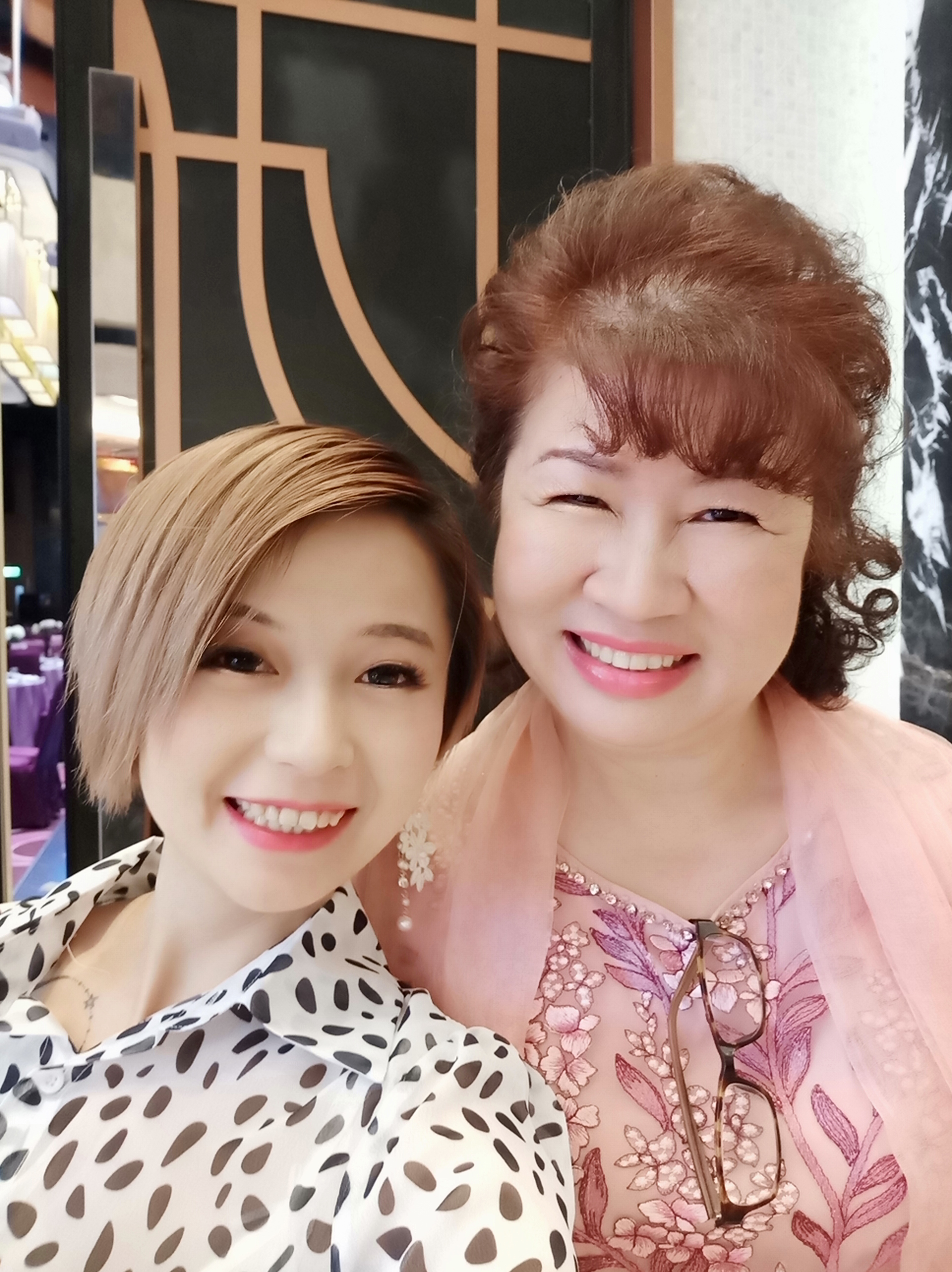 這張照片是兩位女性的合影，她們都面帶笑容，看起來很親密。左邊的女性穿著白色點點襯衫，右邊的女性穿著粉紅色的花朵圖案連衣裙。她們的背景是一面牆，牆上有金色的裝飾。
