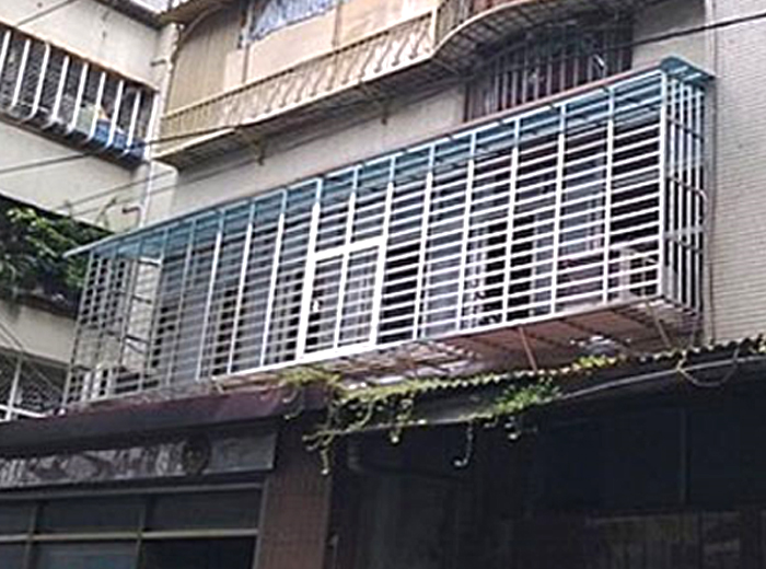 這張照片是一棟建築物，建築物上有一個鐵製的陽臺，陽臺上面有鐵窗，鐵窗是藍色的，陽臺上有一層木板。