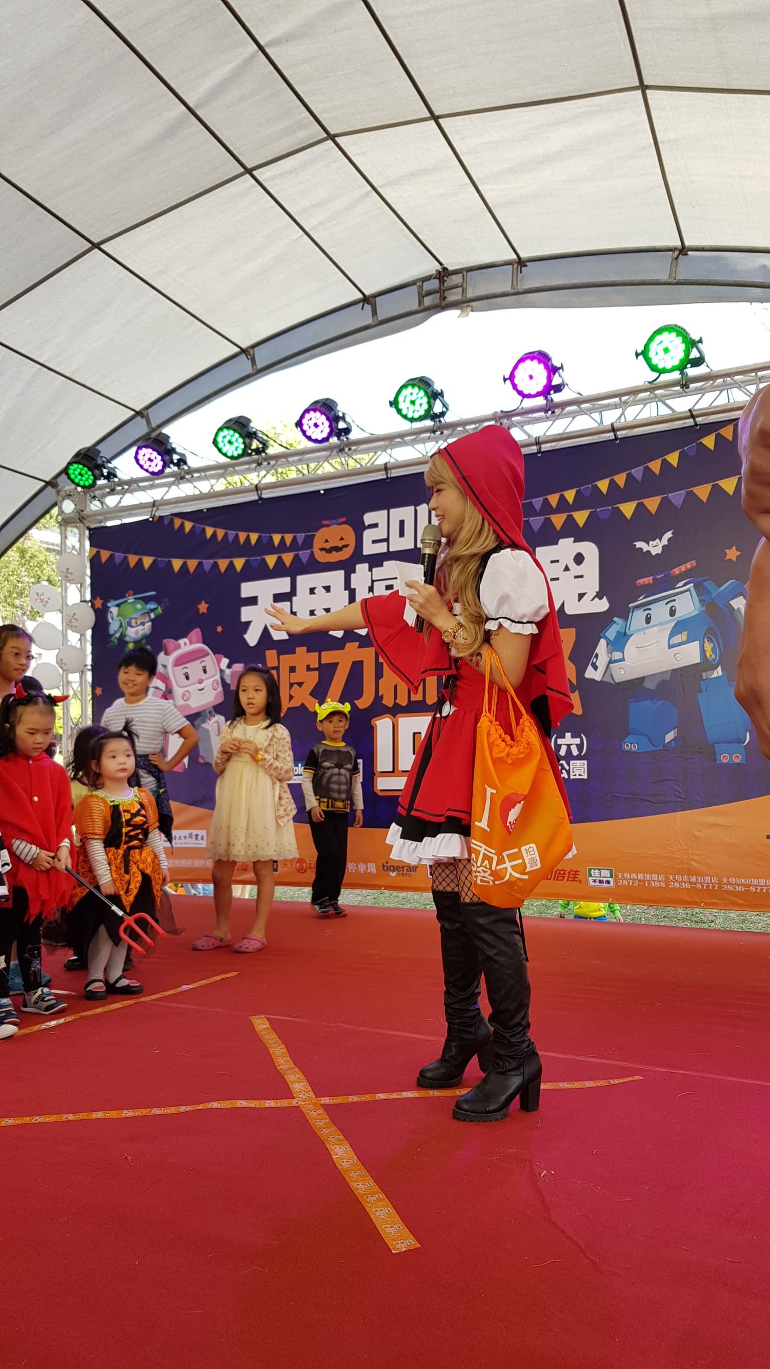 這張「萬聖節活動」的照片是一個身穿紅衣的女孩站在舞臺上，她手持一個橘色的袋子，並且面向著臺下的孩子們。