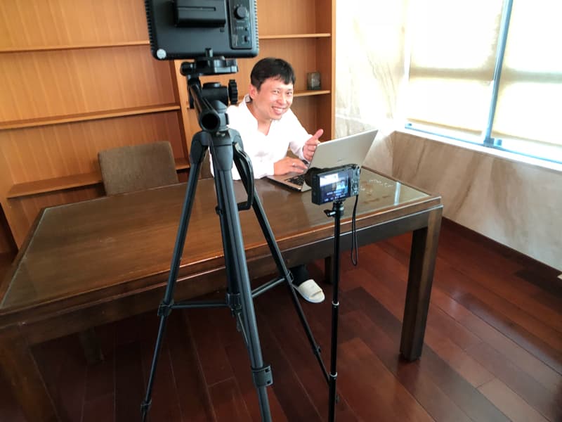 這張「男人正在用電腦錄影」的照片是一個人坐在桌子前，用電腦錄影，他面前放著一臺相機和一個燈光設備。