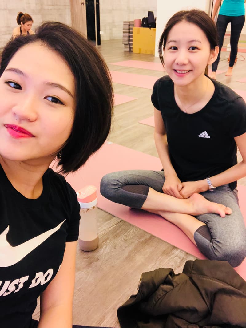 這張照片是一張兩個女生在瑜伽教室的合照，其中一個女生坐在瑜伽墊上，另一個女生站在她旁邊，她們都穿著運動服，看起來都很開心。