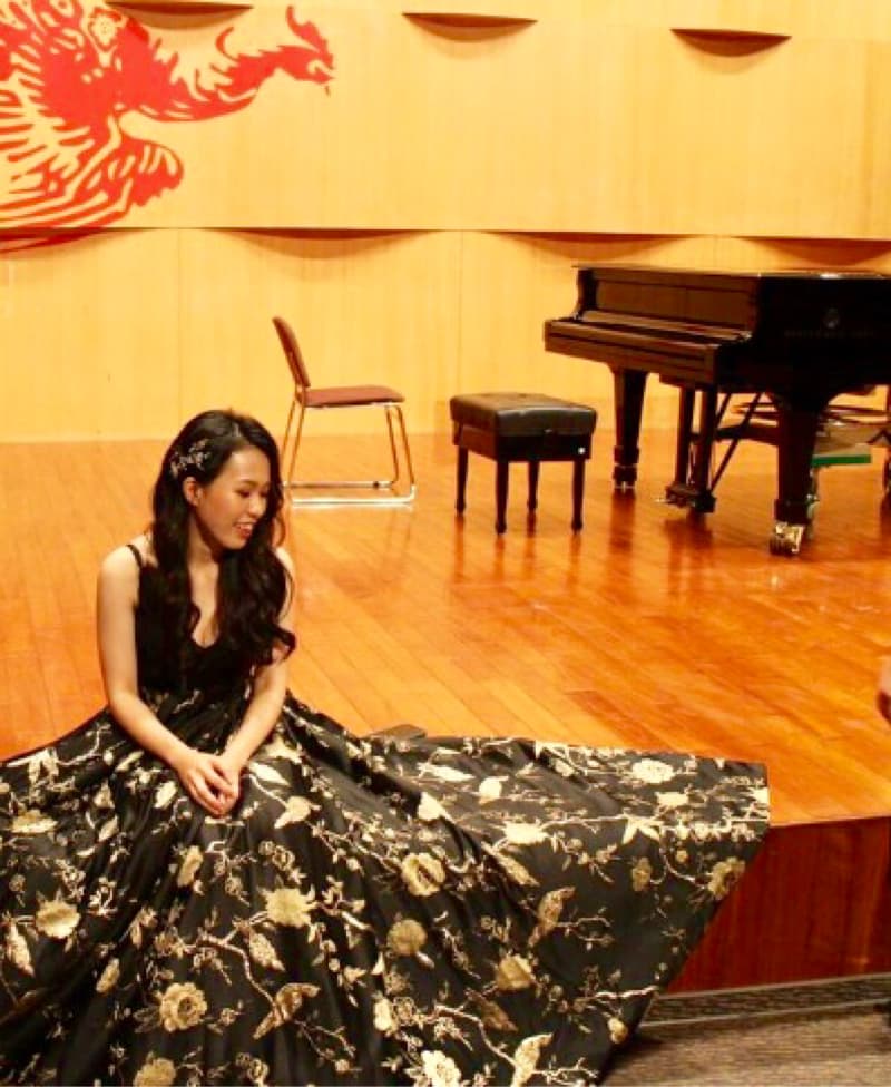 這張照片是一名身穿黑色金線刺繡長裙的女子坐在舞臺上，她的裙子很長，幾乎遮住了整個舞臺。