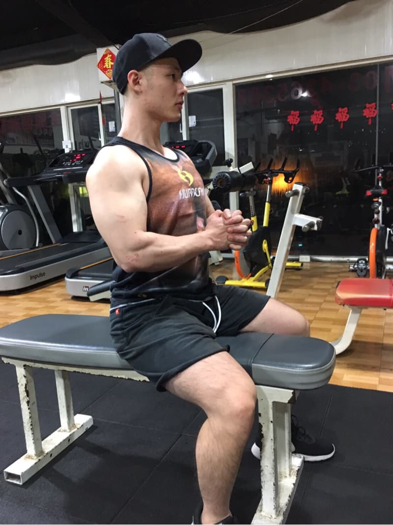 這張照片是一個健身房裡，一個年輕人坐在健身器材上的畫面，他看起來很健壯，正在專注地鍛鍊著。