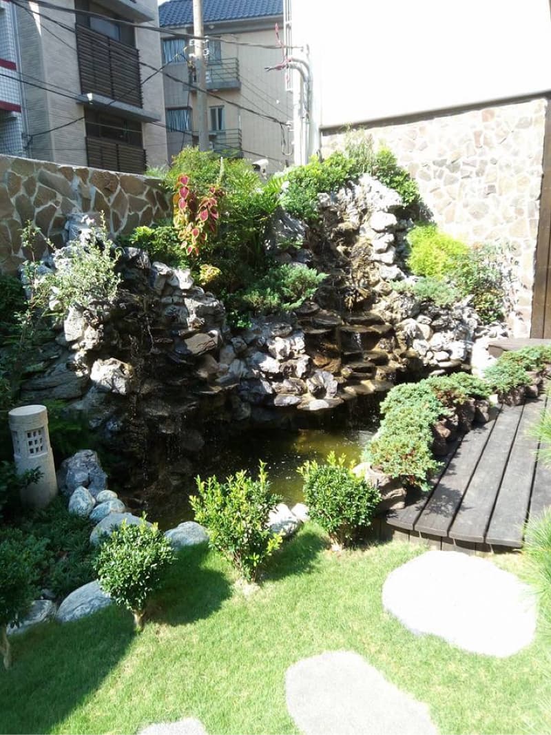 這張照片是庭院裡的人工瀑布，瀑布由許多石頭堆疊而成，水流從石頭縫隙中流下，形成一個小型的池塘，周圍種植著綠色植物，看起來十分寧靜祥和。