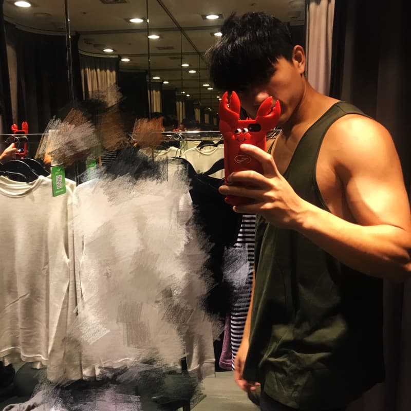 這張「試衣間」的照片是：一位穿著深綠色背心的年輕男子，手持紅色螃蟹造型手機殼在鏡子前自拍，背景是衣架上的衣服。