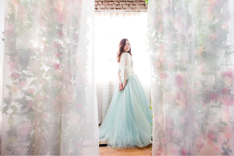 這張照片是一個穿著淺藍色蓬蓬裙的女子，她站在一個佈滿花朵的窗簾前，眼神看向右邊，顯得十分優雅。