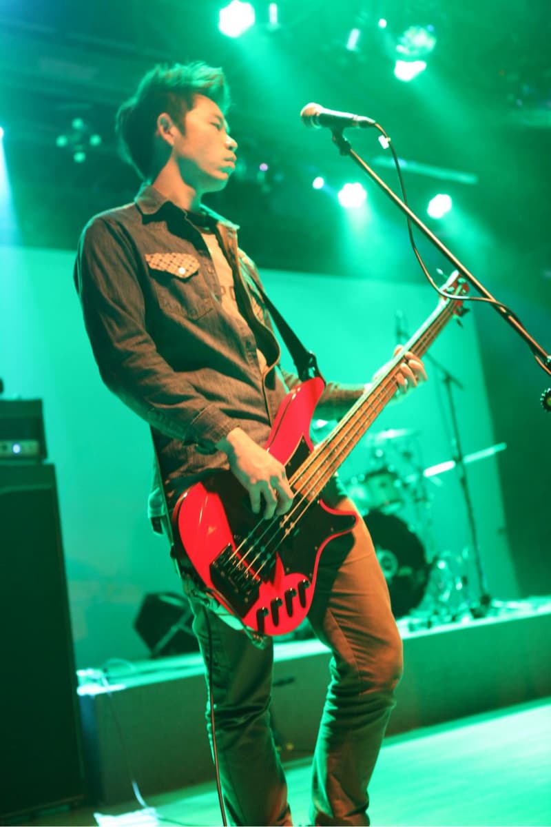 這張照片是一個吉他手在舞台上演奏低音吉他。他穿著牛仔襯衫和卡其褲，正在專注地演奏紅色低音吉他。綠色的舞台燈光照亮了他和他的樂器，給人一種熱鬧的現場音樂表演的氛圍。