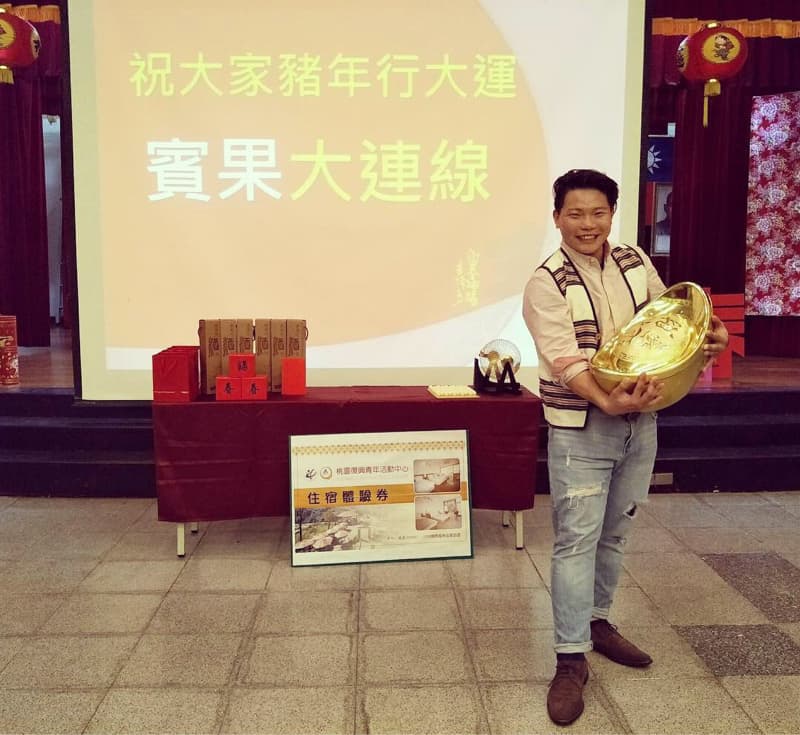 這張照片是一個男子手拿金元寶，站在舞臺上，身後是「祝大家新年行大運，富貴大連線」的布條。