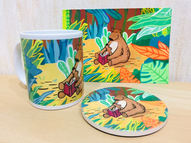 這張照片是一組印有熊親子閱讀圖案的馬克杯和杯墊。馬克杯和杯墊都以熱帶叢林為背景，並以明亮鮮豔的色彩繪製。圖案中的熊媽媽和熊寶寶一起坐在樹葉上，閱讀一本紅色封面的書，畫面溫馨可愛。