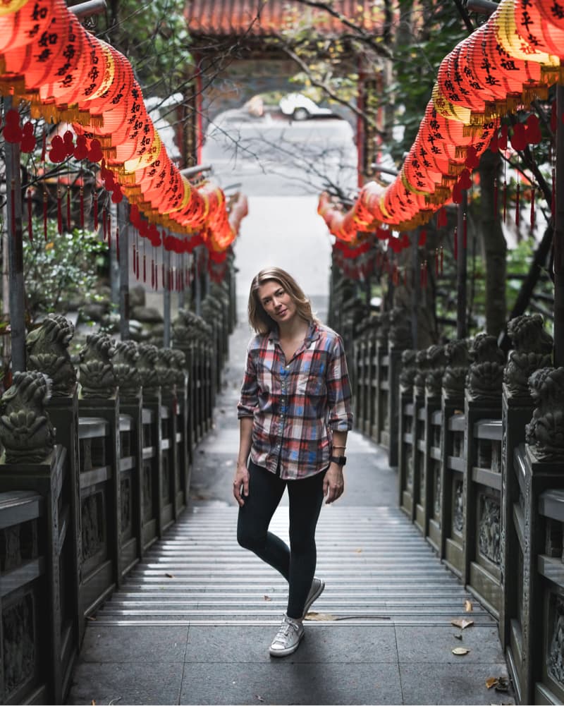 這張「寺廟」的照片是一個年輕女子站在石階上，兩旁是紅色燈籠裝飾的走廊，背景是綠色的樹木。