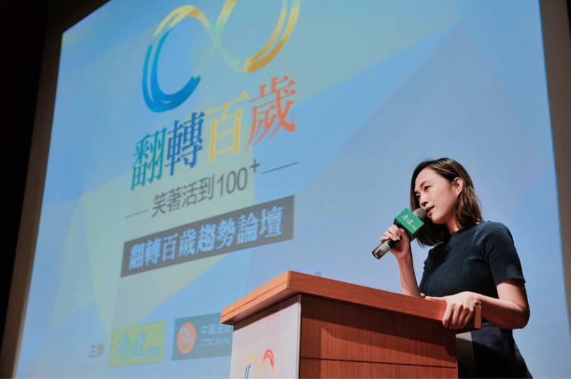 這張「演講」的照片是一個女性站在講臺上，正在用麥克風演講。她身穿深藍色衣服，背景是一塊投影幕，上面印著「樂齡活到100+」的標語。