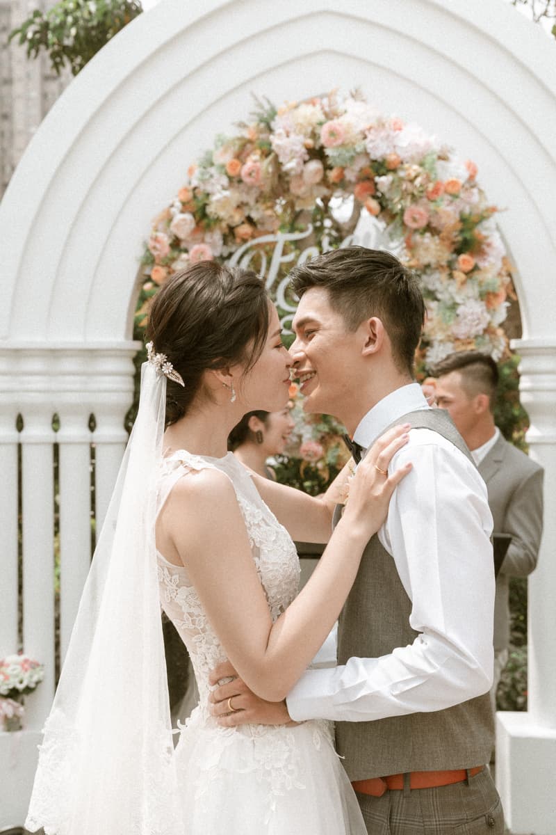 這張照片是一對新人正在親吻，他們穿著婚紗和西裝，背景是一座花拱門。