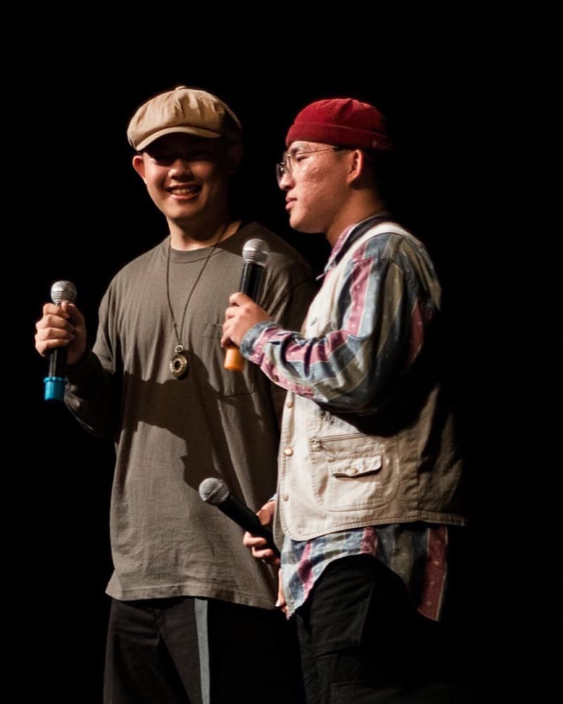 這張「兩個年輕男子手持麥克風站在舞臺上的」照片是，其中一個男子穿著灰色長袖T恤和黑色長褲，另一個男子穿著紅色帽子、白色背心、格子襯衫和黑色長褲。