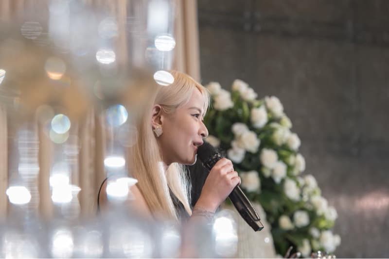 這張照片是一名女性在一個場合中，拿著麥克風，準備演講。她身穿黑色禮服，金色的頭髮散落在肩上，看起來非常優雅。在她面前是一束白色的花，為畫面增添了一絲浪漫的氣息。