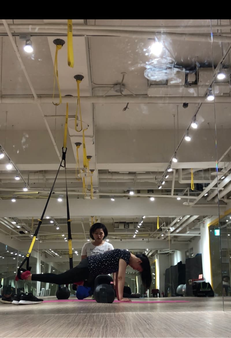 這張照片是一個健身房，一位女性正在進行懸吊式訓練，一位教練在一旁指導。