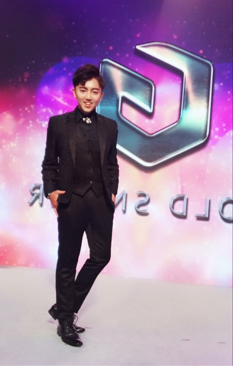 這張「一個穿著西裝的年輕人站在舞臺上」的照片是，他穿著黑色的西裝，打著領帶，站在舞臺上，背景是一個閃閃發光的舞臺。