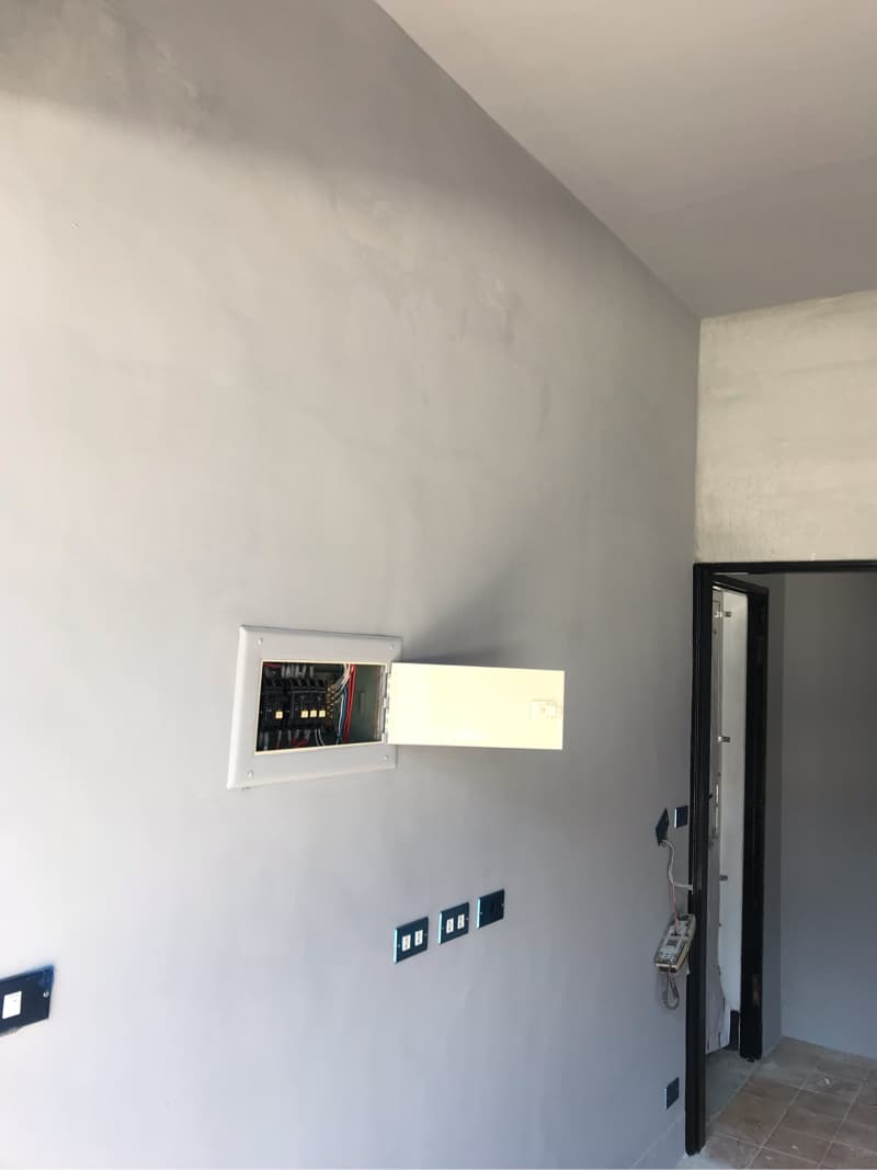 這張「房間牆壁」的照片是，牆上有一個電箱，電箱門打開了，露出裡面的電線和電路。牆上還有幾個插座，旁邊有一扇門。