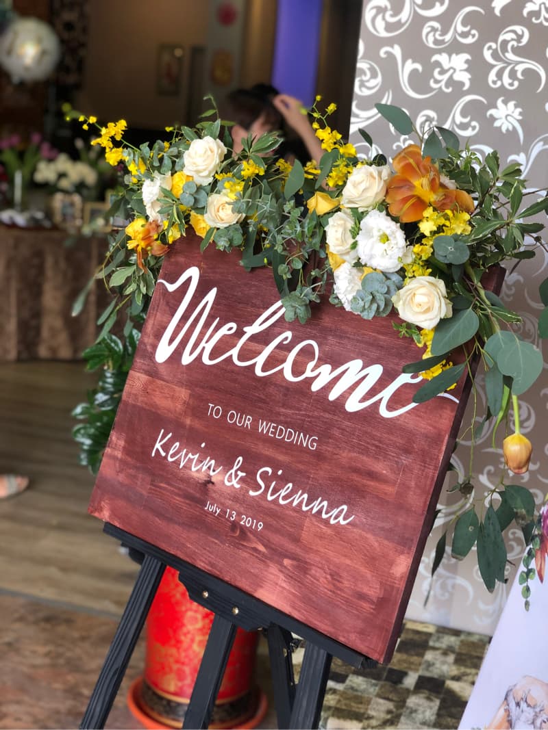 這張「婚禮歡迎牌」的照片是一塊木頭製的牌子，上面寫著「Welcome to our Wedding」，以及新人的名字「Kevin & Sienna」和婚禮日期。牌子被擺放在一個黑色的畫架上，前面擺滿了鮮花。