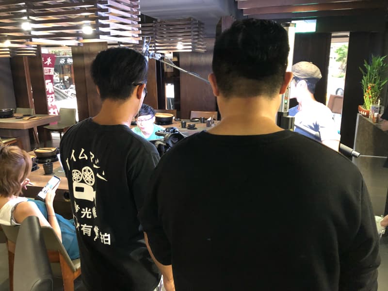 這張「拍攝」的照片是三個人在餐廳裡，其中兩個人站在一起，一個人坐在椅子上，他們好像正在拍攝節目。