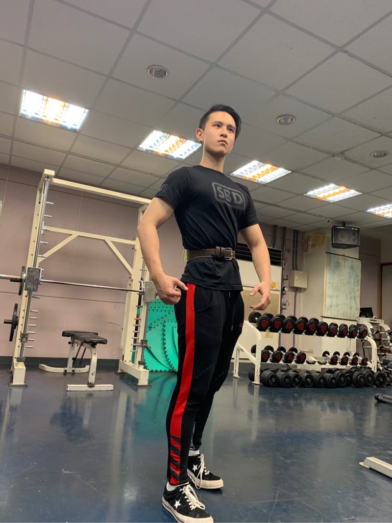 這張「健身房」的照片是一個年輕男子在做健身運動，他穿著黑色運動褲和黑色T恤，他的T恤上有「SBD」的字樣，他站在一個健身器材旁，他正準備做重量訓練。