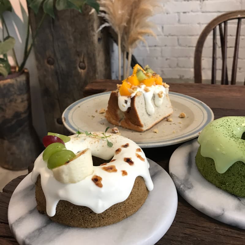 這張照片是一張擺滿甜點的餐桌。桌上有三種甜點，分別是芒果蛋糕、香蕉蛋糕和抹茶蛋糕。最顯眼的是中間的香蕉蛋糕，上面覆蓋著厚厚的鮮奶油，配上新鮮的香蕉和葡萄，看起來美味可口。