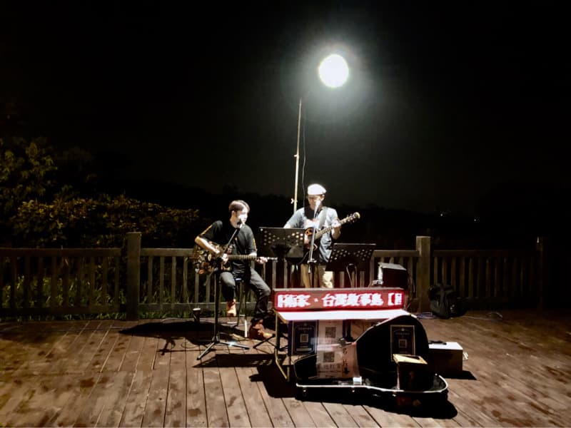 這張照片是一個在夜晚戶外表演的街頭藝人,他們站在一個木製平台上,演奏著吉他,台前放著音箱,旁邊還有他們的收款箱。