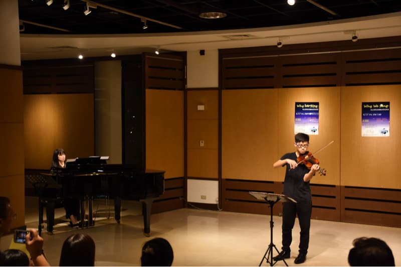 這張照片是一個年輕男子在演奏小提琴，他穿著黑色的衣服，戴著眼鏡，專注地拉著小提琴。舞台上也有一架鋼琴，一位女子正在彈奏。