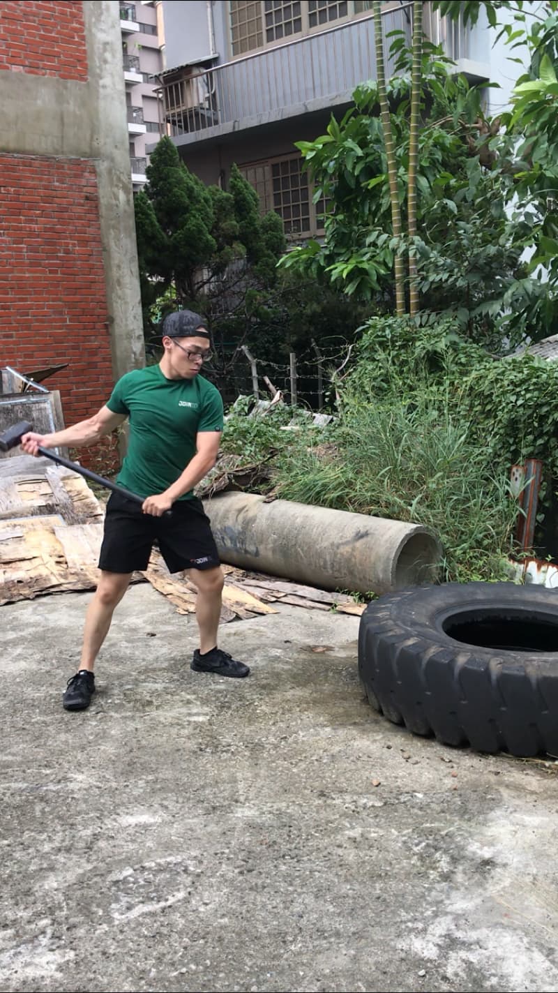 這張照片是一個男人正在用鐵鎚敲打輪胎。他穿著綠色的短袖 T 恤和黑色的短褲，戴著帽子和眼鏡。他正在用鐵鎚敲打一個黑色的輪胎，輪胎放在水泥地上。