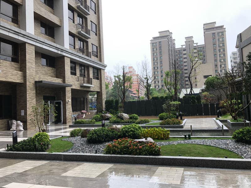 這張照片是一棟現代化公寓大樓的建築外觀，最重要的是公寓大樓前方的庭園，庭園設計優雅，種植了許多綠色植物，並鋪設了石頭路和石頭造景，營造出寧靜舒適的氛圍。