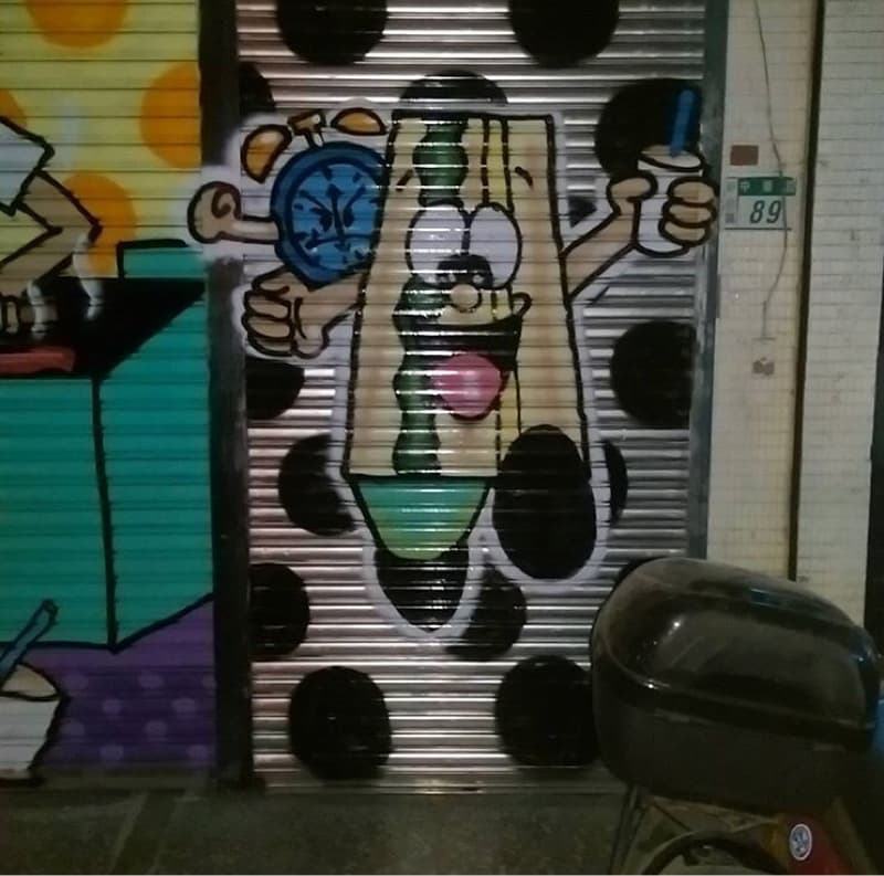 這張「塗鴉」的照片是一個穿著三明治服裝的人，他手裡拿著一杯飲料，背景是帶有黑色圓點的灰色捲簾。