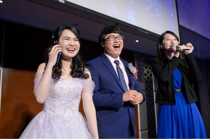 這張照片是一對新人正在婚禮上接受祝福，新郎穿著深藍色西裝，新娘穿著淡紫色禮服，兩人笑容滿面，看起來非常幸福。