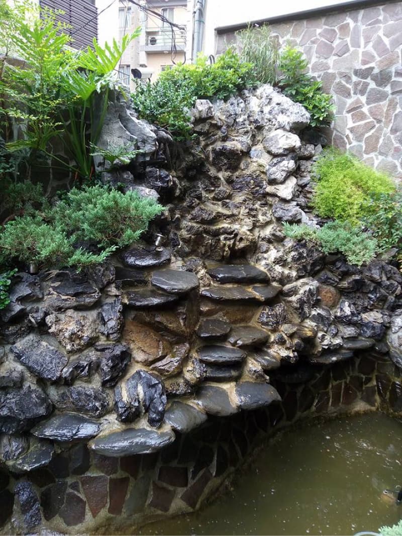 這張照片是一座用石頭堆砌的小型瀑布，周圍環繞著綠色的植物，呈現出自然寧靜的氛圍。