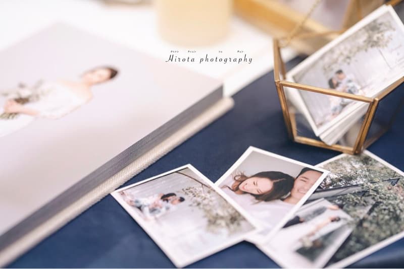 這張照片是幾張婚禮照片，包括一本相簿，一個透明玻璃盒子裡有幾張照片，以及幾張散落在藍色桌布上的照片。