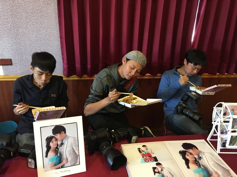 這張「婚禮攝影師在餐廳吃飯」的照片是三個攝影師在婚禮現場餐廳吃飯，桌上擺著婚禮照片、相機、閃光燈等。