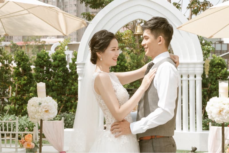 這張「婚禮」的照片是一對新人在白色拱門下互相凝視，新娘穿著白色婚紗，新郎穿著白色襯衫和灰色背心，他們看起來非常幸福。